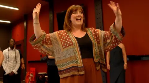 Marie Claire Breen, vocal coach at Opera North in bright patterned red and mustard jacket with black top and arms up, going through vocal warm up before the rehearsal