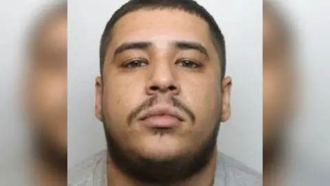 A police mugshot of Lakan Illsley. He has dark hair and a dark beard and is wearing a grey T-shirt. He is stood against a light-coloured background.