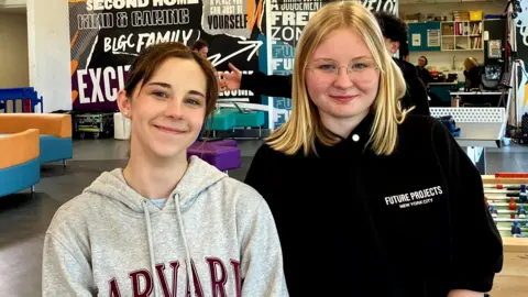Two girls smiling at the camera as they pose for a picture in a youth club. Maisie on the left has brown hair pulled back in a pony tail and is wearing a grey hoodie with Harvard University branding in large red type. Iga on the right wears a black hoodie bunched up on her arms. She has shoulder-length blond hair and has rimless glasses.