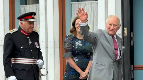 Becki Bowden/BBC King Charles dressed in a grey suit and red tie waving to onlookers as he arrives at the CARE Hub in the town.