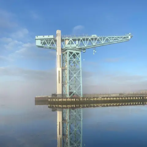 Douglas King A large, light green crane stands on a dark pier jutting out into still water. The water is reflecting the blue sky above, which is streaked with white cloud. There is grey cloud in the background.