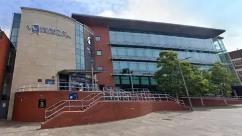 The university building is glass and brick and has steps leading up to the entrance with trees on the right-hand side.