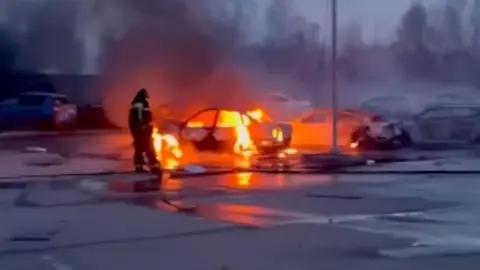 Man stands next to burning car in Moscow. The shell of the car is glowing red.