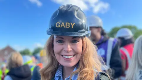 Amanda White/BBC A woman with long white-coloured hair smiling at the camera. She is wearing a black hard hat labelled 'GABY' and a striped shirt under a black jacket. People are gathered in the background. The sky is blue.