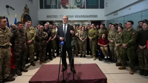 Reuters Sir Keir Starmer mid-speech wearing a dark suit, standing on a podium with two microphones, surrounded by dozens of men and women in military uniform.