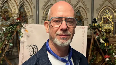 Choir director Rónán de Búrca pictured at the chapel. He is wearing a denim jacket and a blue neck-tie and is smiling into the camera. He has a bald head with some white hair on the sides and a white and grey beard. He is also wearing glasses.
