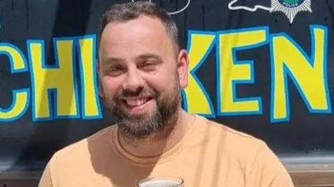 Staffordshire Police A man with a dark brown beard and short dark hair smiles showing his teeth to the camera. He wears a light orange top and appears to have a beer glass in front of his chest. Behind him are yellow letters with a blue outline spelling "chicken"