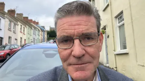 Shows a man wearing glasses with a light blue jacket standing in a street with cars in the background.