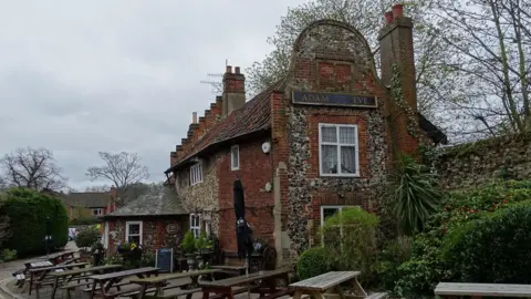 Matthew Chadwick/Geograph The Dutch gable end of a red brick and flint pub, with the sign Adam and Eve above its second floor window. There are pub benches outside.