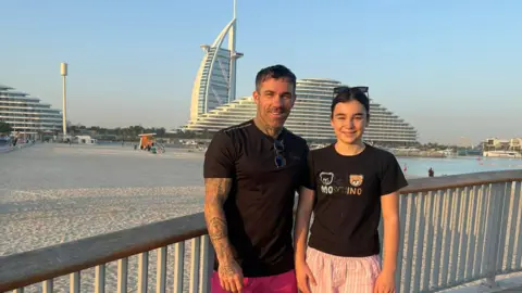 Ryan Hodgson stands ext to his daughter in front of The Palm Dubai. They are both wearing black tshirts. There is a beach behind them and blue skies. 