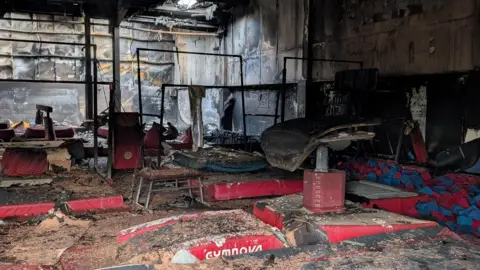 Lynx Gymnastics Centre The inside of a gym, showing equipment burnt, with lots of ash. All the items are burnt and part of a roof has collapsed with light coming in.