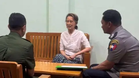 Myanmar state TV Aung San Suu Kyi, an elderly woman sitting on a bench with her arm on the rest. In the foreground two uniformed men sit either side of her at a table.