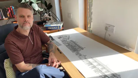 A man with grey and brown hair slicked away from his forehead is sat in a chair next to a desk looking at the camera. He has his hands together and knee crossed and is wearing a brown short-sleeved shirt and blue trousers. On the table is a long paper roll with detailed drawings of buildings.
