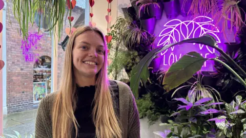 A young woman with long blonde hair is pictured wearing a green woolly cardigan while sat in a shop window. Behind her are several plants and leaves with a large illuminated purple sign that reads "LOV LEAF". 