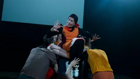 Luca Truffarelli Joanna Banks is held up by a group of dancers. She is wearing orange clothing and a black and orange headscarf and white trainers. A screen is behind her.