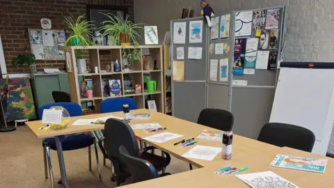 Wayfinder Woman A room with tables set up for activities, with chairs, paper and pens, shelves with plants and books, and display boards with posters.