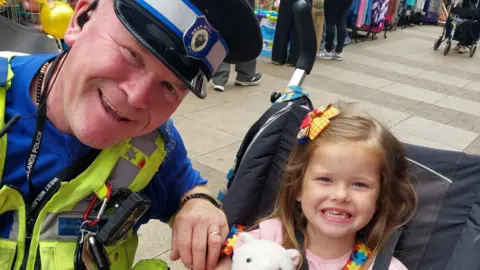 A smiling man in a PCSO uniform of a blue top with hi-viz vest on top with a radio and keys attached, and a black cap with blue and white hatband. He is crouching down by a smiling toddler in a pushchair with shoulder-length brown hair and a yellow-and-red hair grip.