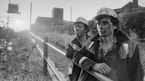 Two young men stand with a setting sun against their backs. They have dirty faces and are wearing worker's clothes.