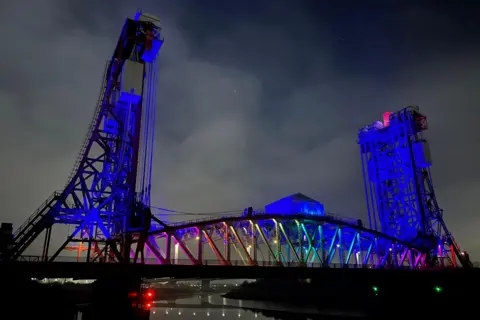 BBC Weather Watchers / Tony Winward The metal Newport Bridge over the River Tees is lit in blue at the edges with rainbow lights illuminating the middle. There are metal lifts on either side of the bridge which rise into the air. The metal structure connecting the two-side is an arch. Clouds can be made out in the dark night sky.