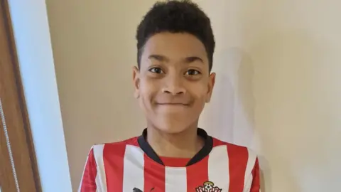 Nicky Ajanaku Kayo Ajanaku - A young boy wearing a Southampton red and white striped football shirt. He is looking straight at the camera and is smiling. He is stood in front of a plain white wall and a solid wooden door. 
