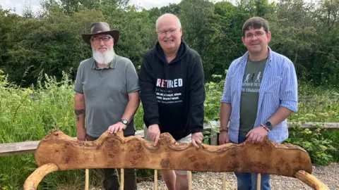 Three men standing outdoors behind a decorative wooden bench. The first man has a bushy white beard and a brown wide-brimmed hat, the second has white hair, glasses and a dark top, the third has brown hair, glasses and a blue striped shirt over a green tshirt