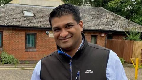 West Suffolk Council  A man stands outside a building in a car park and smiles at the camera. He has short dark hair and is wearing a black gilet jacket with a blue shirt underneath. 