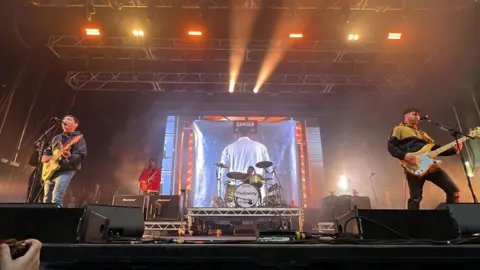 BBC/ Marian McNamee The Enemy perform on the main stage at the Godiva Festival in 2023. At the front of the stage are two men playing guitars in front of microphones.