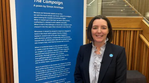 Johnny Reed / BBC A woman with a brunette bob smiles. She wears a ruffled shirt and a black blazer. Beside her is a 6 foot print of the poem