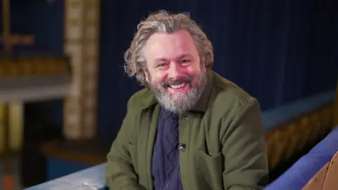 BBC Michael Sheen, yang memiliki rambut panjang beruban dan berjanggut, tersenyum ke arah kamera sambil duduk di teater 