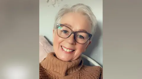 Patricia Daglish smiling at the camera. She has short white hair, blue eyes and glasses. She is wearing a brown jumper.