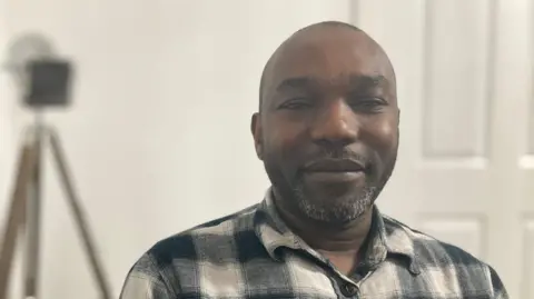 BBC Dad of two Ambi Ambituuni is sitting on his sofa at home and is wearing a plaid shirt