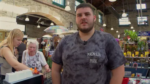 Kieron Loomes, 26, standing at his market stall in Tavistock pannier market wearing a grey t-shirt with customers behind him looking at his products