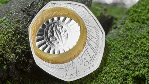 A single coin on a fake background of granite grey rock frosted in green lichen. the coin is silver with an etched inscription in Elfish, a Lord of the Rings script and language. A gold band, the one ring, also inscribed in Elvish, is at the top left of the seven sided coin. the ring surrounds the eye of sauron etched onto the silver coin with an oval around the eye which is surrounded by a fan of lines to the ring. 