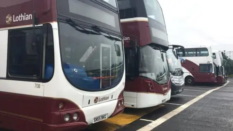 Several maroon and white double-decker Lothian buses parked in a row at a bus depot on a wet day, with the nearest bus showing registration SN55 BKG.