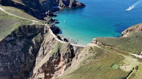 BBC Aerial shot of La Coupée, a narrow road in Sark which connects two parts of the island. The road has a rockface below it. A boat can be seen heading towards the island in the top right-hand corner of the image.