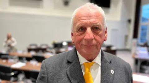 Simon Dedman/BBC Barry Aspinell wears a grey suit, yellow tie and white shirt. He has white hair. He looks just off camera, with a council chamber out of focus behind him.