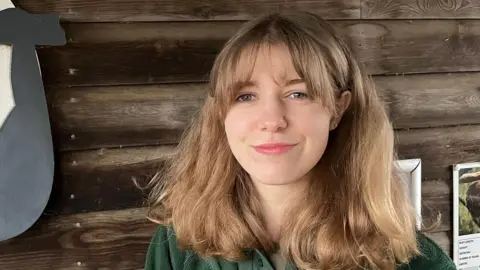 John Devine/BBC Megan Jones is standing off centre to the right. She has shoulder length dark blonde hair and fringe and is wearing a green T-shirt and jacket. She is standing in front of a wooden wall.