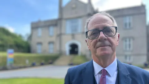 Emlyn Dole looks at the camera, with the university blurred in the background. He has short silver hair and glasses, and is wearing a blue suit jacket, white shirt and red checked tie.