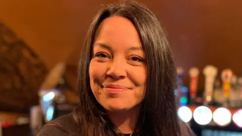 BBC/Lucinda Adam A woman with long, black hair wearing a black jumper and standing in front of a beer tap.