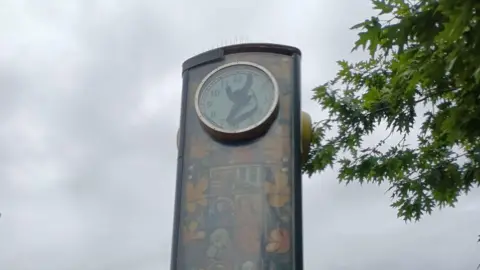 Image of The Keynsham Clock. It is a tower, with many small, square images on its base. The clock face is covered in a thick, black material. One of the clock hands can be seen broken off and lying across the edge of the clock, between the numbers 7 and 9.