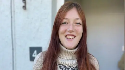 A teenage girl is standing in front of the door to her home. She is wearing a turtle neck jumper which has a butterfly on the front at the top, and along her lower arms. She has long dyed brown hair, and a big smile.