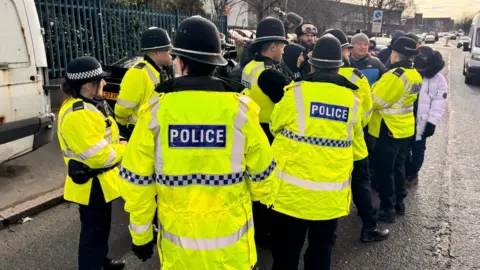 Unite Police officers crowing around protesters on a street. They wear high-vis green jackets with the word police in white letters on the back. Most of the officers wear helmets. There is a group of protesters beyond the officers.