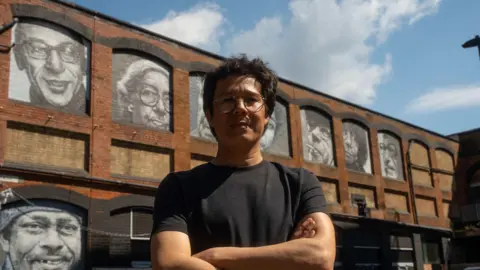 Benji Mitchell, Dancing Light Films A general view of Abraham O, a London-based street artist, looking at the camera with his arms folded. In the background, there are seven murals that he made with spray paint. One is of Emanuel Litvinoff.
