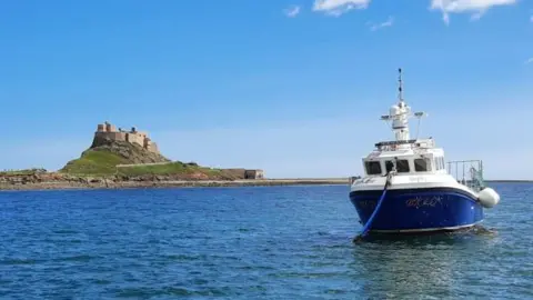 NIFCA A small fishing boat with a blue hull and white cab and deck is moored up off Holy Island, with a castle on a hill in the background