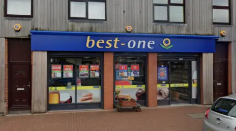 A shop with the words Best One on a blue background at the top of the ground floor outside. Part of a pavement and a silver car are visible in front of the store.