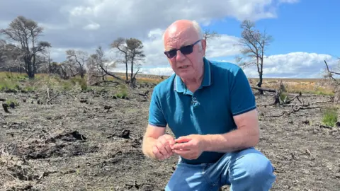 Lol Hodgson is kneeling on blackened moorland. He is wearing dark sunglasses and a blue t shirt and blue jeans