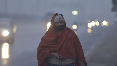 A woman wearing a shawl walks on a road in Uttar Pradesh state, with vehicle lights barely visible in the background because of the dense fog. 