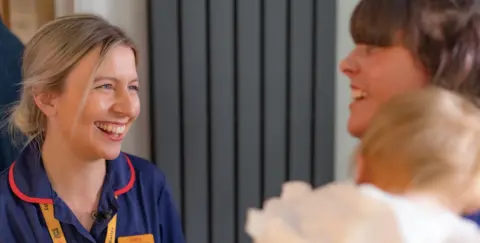 Roald Dahl's Marvellous Children's Charity A woman with blonde hair that is tied back into a bun smiles at another woman with brown hair who is holding a baby with fair hair who are blurred slightly. 