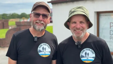 John and Eamon pictured together, both wearing black t-shirts and sporting short grey beards. One wears a green bucket hat and the other a brown peaked cap. 