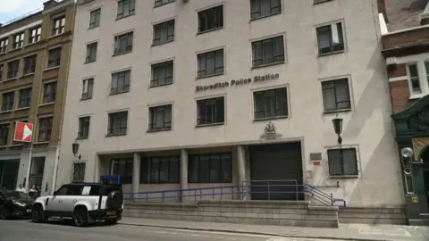 The exterior of Shoreditch police station: a light grey building, with blue railings outside and the shutters pulled down over its entrance